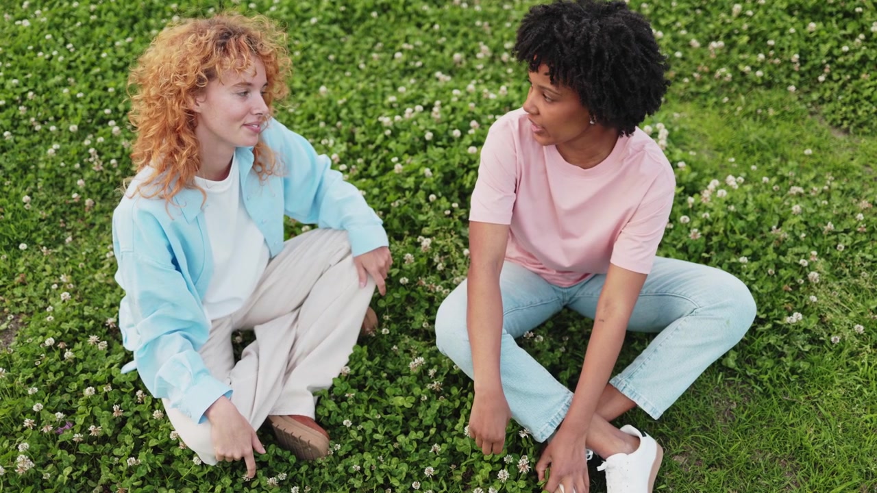 Due giovani donne diverse sedute sull'erba e che parlano felicemente