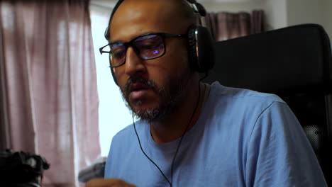 South-Asian-man-with-glasses-and-headphones-air-drumming-at-his-desk-at-home,-engaging-in-music-enjoyment-and-relaxation