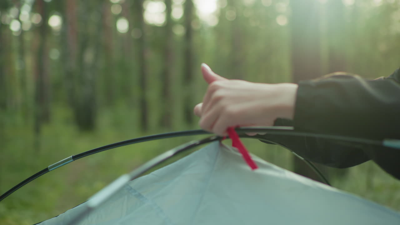 close up hand tying red tent rope around flexible tent pole during peaceful camping morning in forest as soft natural sunlight creates glowing effect through trees and illuminates focused action