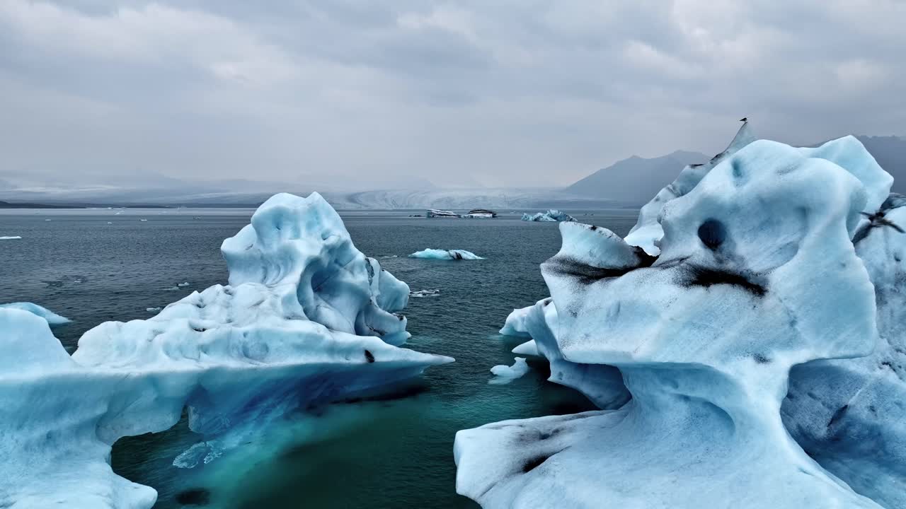 Floating icebergs drift through cold Arctic water, showcasing dramatic blue ice formations and a rugged glacial landscape under overcast northern skies