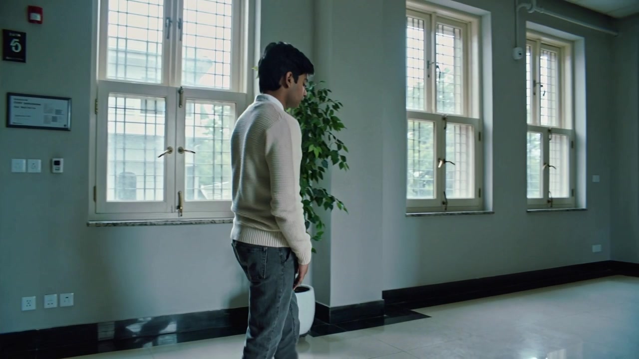 Pausing near double doors, man wearing cream sweater walking past windows and potted plants in hall