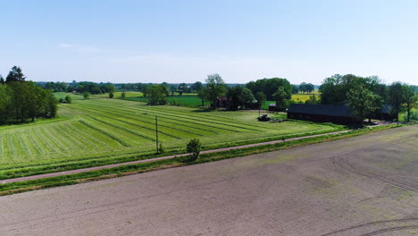 Establishing-drone-clip-of-a-Swedish-summer-field-with-a-tractor-gathering-dried-grass-in-rows-on-a-warm-sunny-day-with-clear-blue-skies-without-clouds-at-a-small-yard