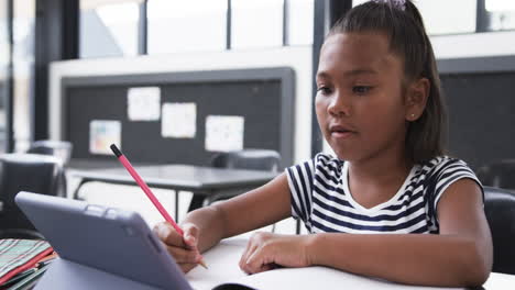 in a school classroom, a young biracial girl focuses on her tablet