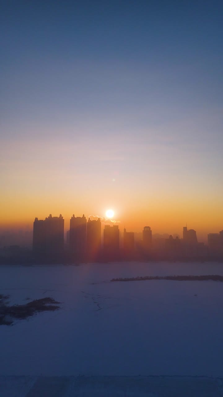 Vertical aerial view of Harbin’s skyline at sunrise, with golden sunlight illuminating the frozen Songhua River, creating a stunning contrast between the icy landscape and the warm morning glow. China