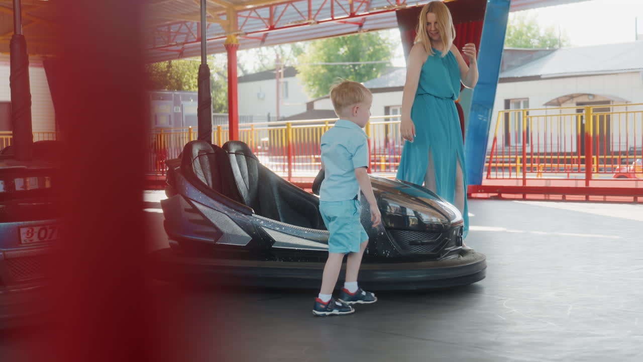 Un bambino che sale su un'autoscontro al parco, una scena di luna park illuminata dal sole con la madre che aiuta il bambino a salire sull'attrazione, un padiglione colorato e un movimento delicato, sorrisi spontanei e legame durante una gita in famiglia nel fine settimana.