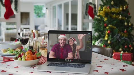 Happy-caucasian-couple-in-santa-hat-on-laptop-lying-on-christmas-table