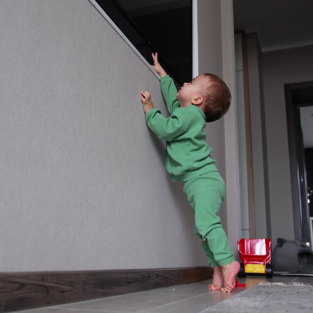 Curious kid tries to reach the TV set in the alcove of the wall. Little toddler stands on his toes holding by the wall