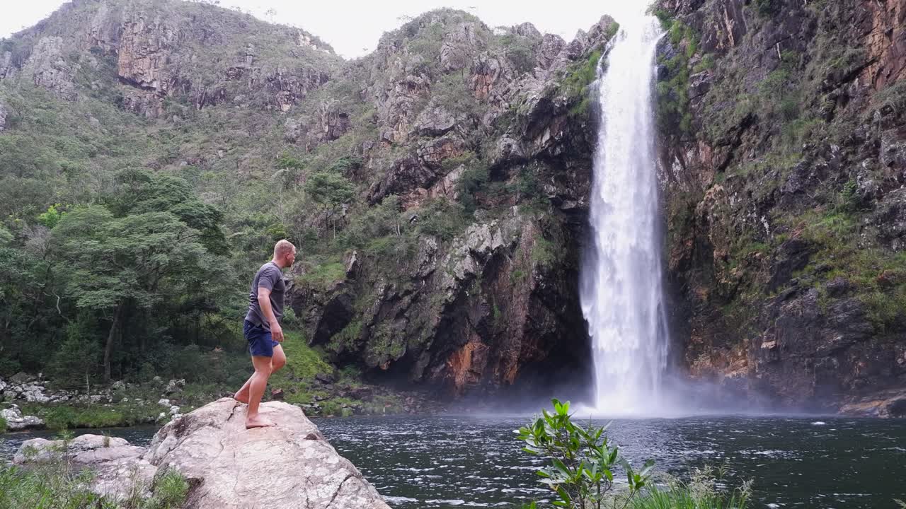 Seated young male tourist rises from waterfall pool and walks to camera