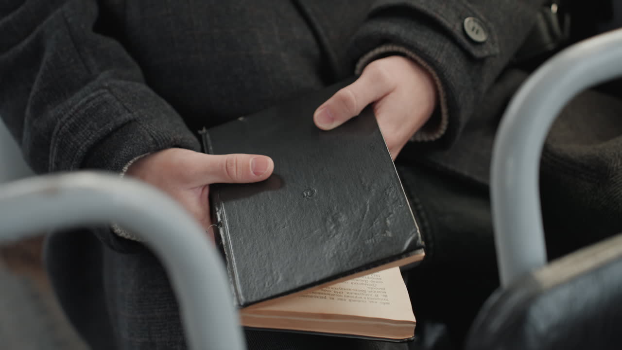 Hænder holder sort notesbog i bus, skaber skriver dagbog under pendling, ærme af uldfrakke, læderomslag og gamle sider titter frem, metalsædeskinne indrammer nærbillede, dæmpede farver og eftertænksom stemning.