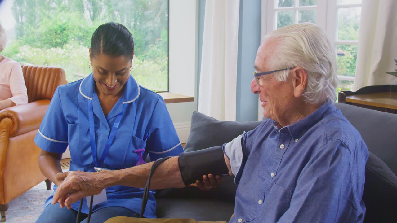 Senior Man At Home In Lounge Having Blood Pressure Taken By Female Care Worker In Uniform