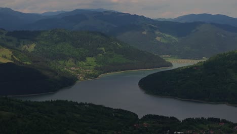 el primer plano aéreo muestra un lago de acumulación rodeado de montañas verdes y boscosas, parcialmente nubladas.