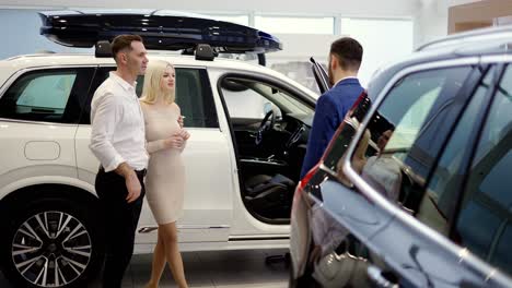 Couple-talking-to-car-dealer-in-dealership-discussing-automobiles-looking-at-luxurious-new-white-model