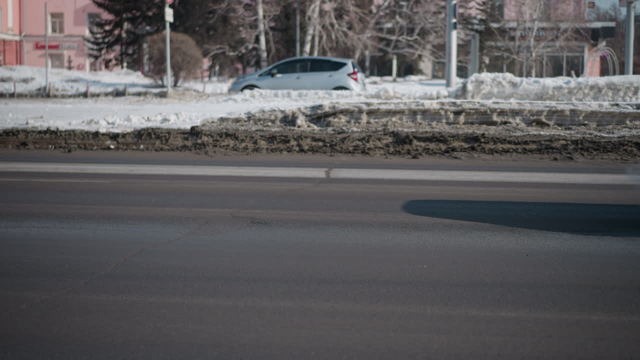 Cars moving along both sides of road past dirty snow median, winter street with speed sign post, trees and pastel buildings beyond, wet asphalt and slush in foreground, bright daylight city