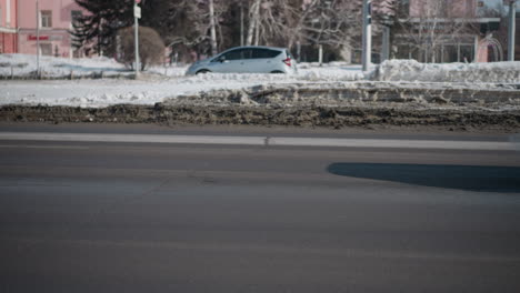 cars moving along both sides of road past dirty snow median, winter street with speed sign post, trees and pastel buildings beyond, wet asphalt and slush in foreground, bright daylight city