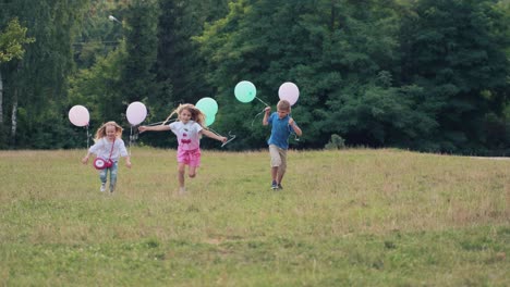 joyful children with balloons in their hands run along the green grass in the park. the boy and girls are having fun