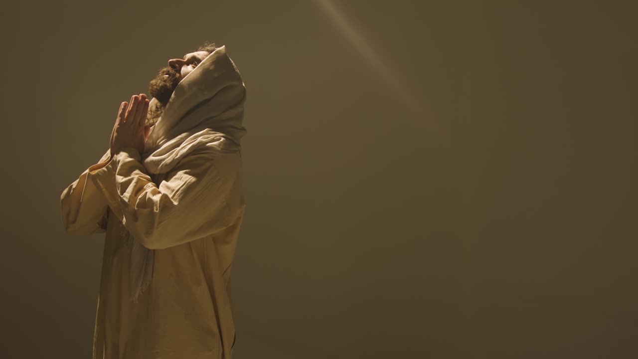 retrato de estudio de tres cuartos de longitud de un hombre vestido con túnicas con cabello largo y barba que representa la figura de jesucristo mirando hacia arriba y orando