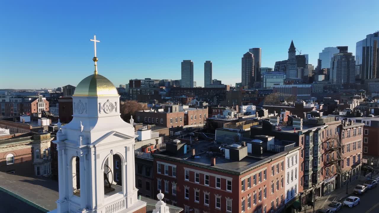 Golden dome crowns St. Leonard’s Church in Boston’s North End. Skyline rises behind brick rowhouses. Aerial.