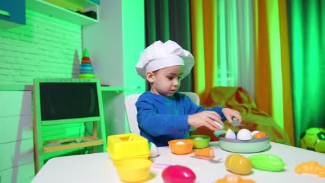 cute caucasian baby boy in white chef cap sits at table. little child playing actively with toy crockery and food.