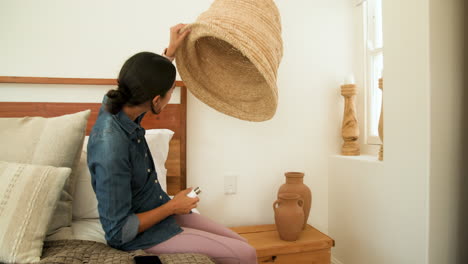 woman adjusting woven lamp shade in cozy bedroom with smartphone on bed, at home