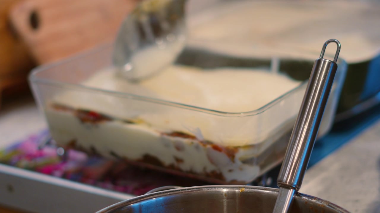 Molho Bechamel sendo espalhado sobre camadas de massa para preparar lasanha em travessa de vidro, pronto para cozinhar em forno quente 4K