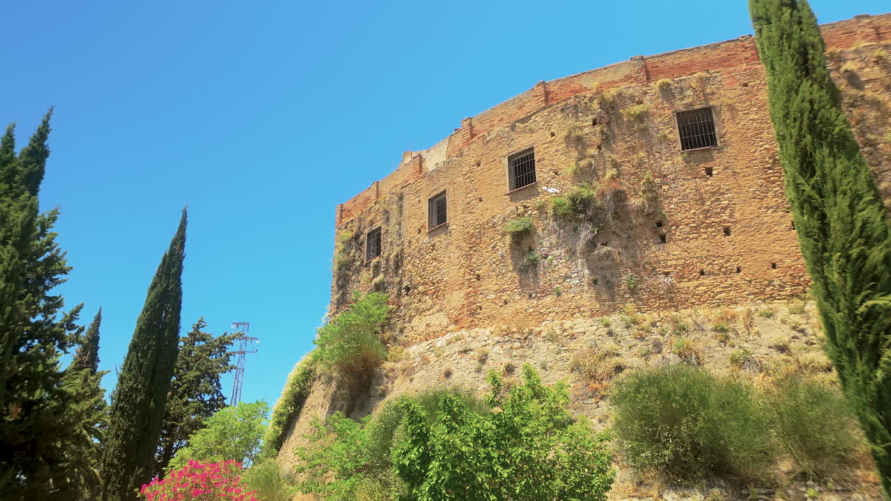Ancient stone wall of a fortress in Ronda, Spain, surrounded by tall cypress trees under a vibrant blue sky. A testament to the town’s history