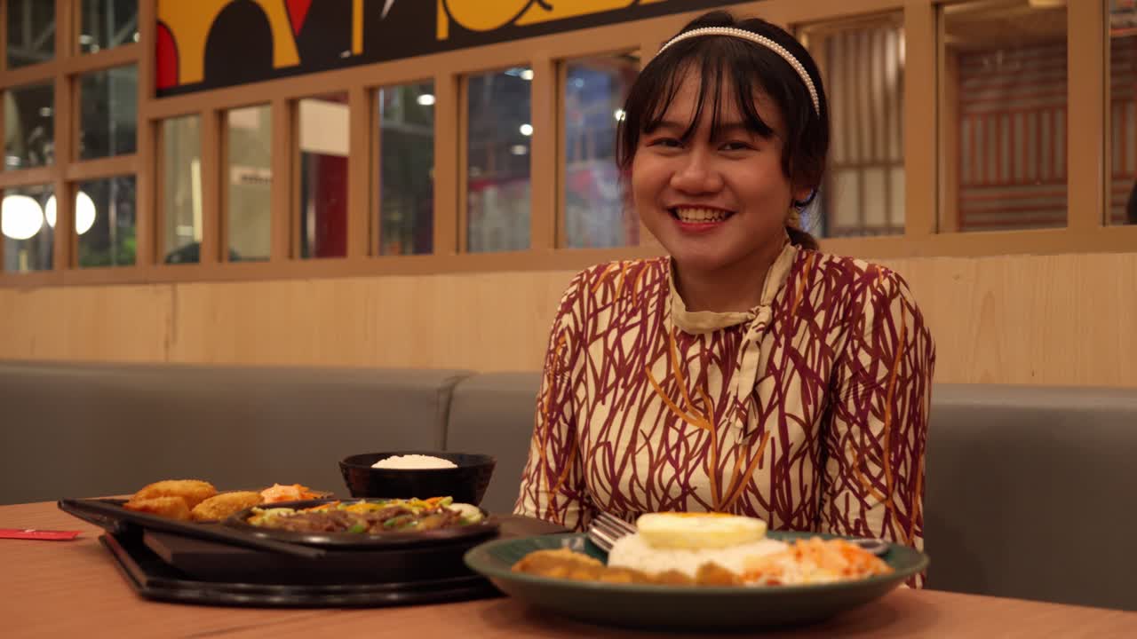 A smiling young woman enjoys Indonesian food at a cozy restaurant with a cheerful atmosphere