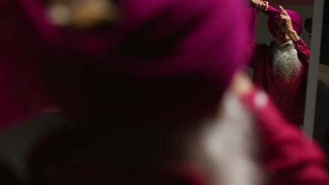 low key studio lighting shot of senior sikh man with beard tying fabric for turban against dark background looking in mirror 2