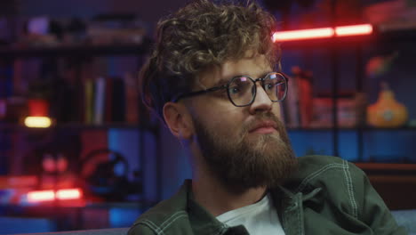 close up view of cheerful young man in glasses and with curly hair smiling to the camera in cozy living room at night