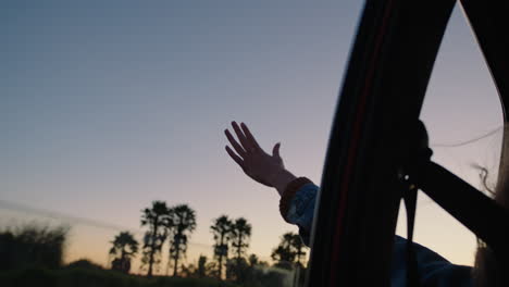 Frau-Im-Auto-Hält-Die-Hand-Aus-Dem-Fenster-Und-Spürt,-Wie-Der-Wind-Durch-Die-Finger-Weht,-Während-Sie-Im-Sommerurlaub-Auf-Dem-Land-Unterwegs-Ist-Und-Bei-Sonnenuntergang-Die-Freiheit-Auf-Der-Straße-Genießt