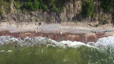 Overhead-drone-views-showcase-the-rugged-charm-of-a-high-cliff-coast-in-Lithuania's-Baltic-Sea,-where-powerful-waves-meet-the-enchanting-green-sea,-painted-with-moody-colors