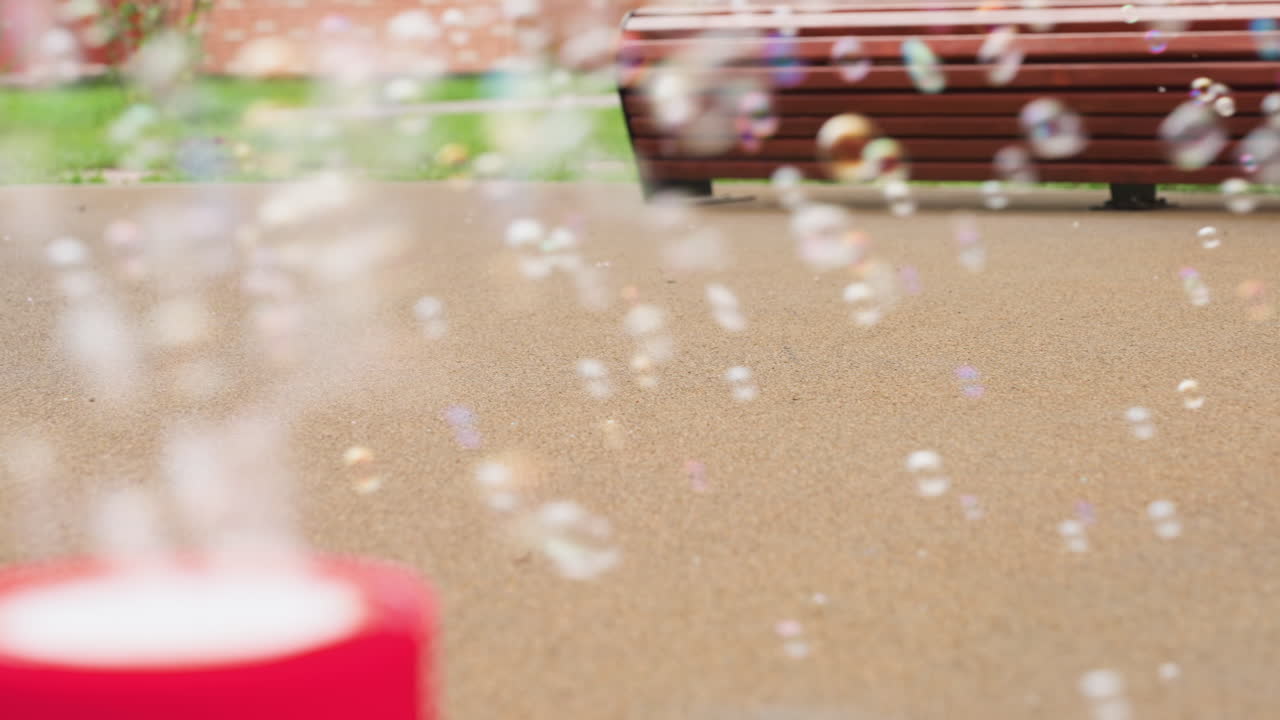 Close up of soap bubbles drifting over pavement, soft bokeh of bench edge and green grass behind, gentle breeze moves tiny spheres across courtyard