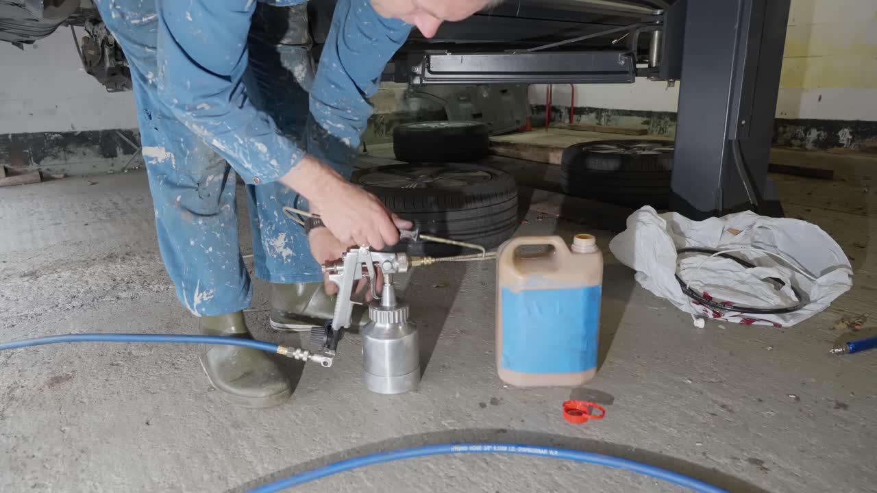 Man prepares to apply lanolin rust prevention spray to car underside, testing spray gun flow with hose and angled nozzle attached