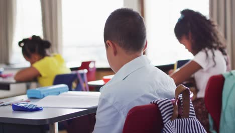 retrato de un niño de escuela de raza mixta feliz sentado en el aula, tomando notas, mirando a la cámara
