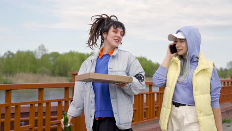 young woman with dreadlocks drinking beer and holding pizza box while her friend talks on the phone