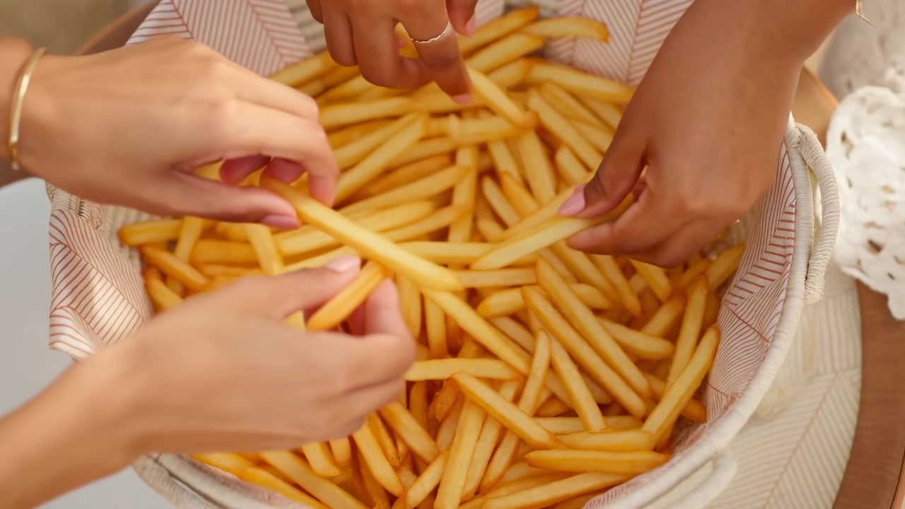 Multiple hands reaching into woven basket of golden fries, sharing warm snack in close-up table scene with linen cloth, soft light, jewelry details, and casual social dining mood