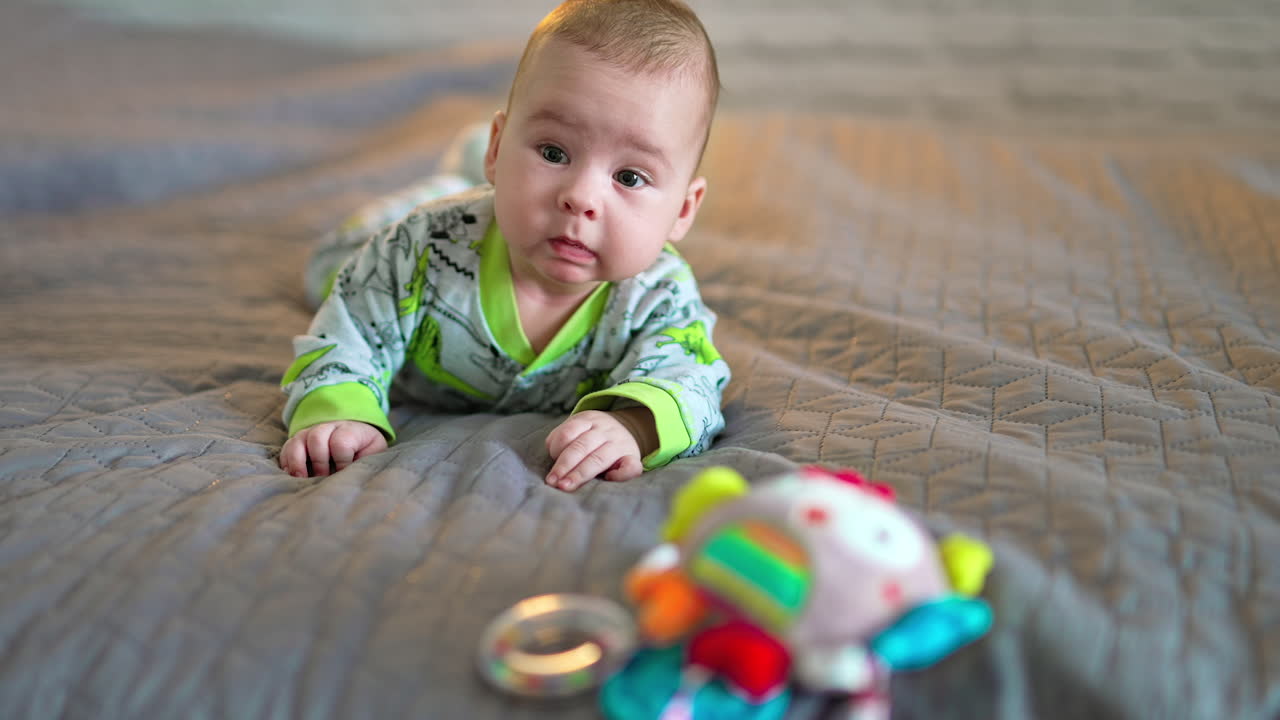Lovely beautiful kid lies on big bed and looks at a bright toy in front of him. Grey background in blur.