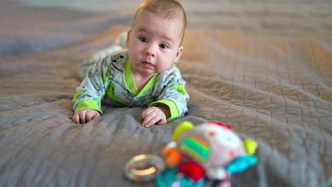 lovely beautiful kid lies on big bed and looks at a bright toy in front of him. grey background in blur.