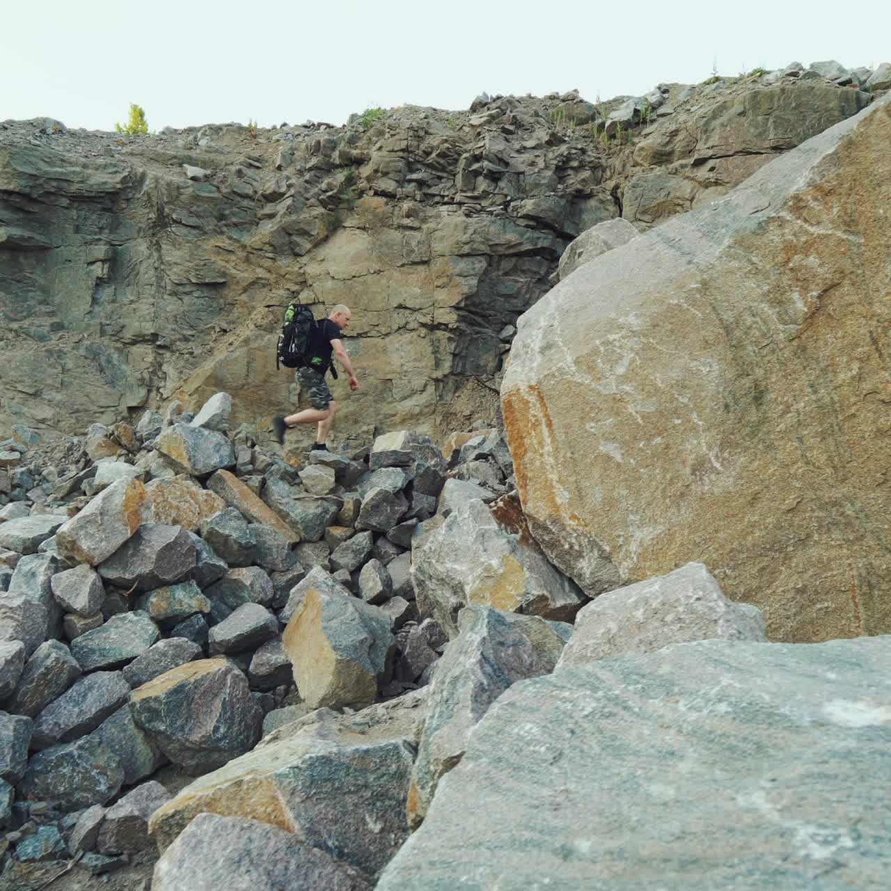 traveler in sportswear with a backpack on his shoulders is going on a pile of stones to a huge stone on the background of rocks in warm summer day