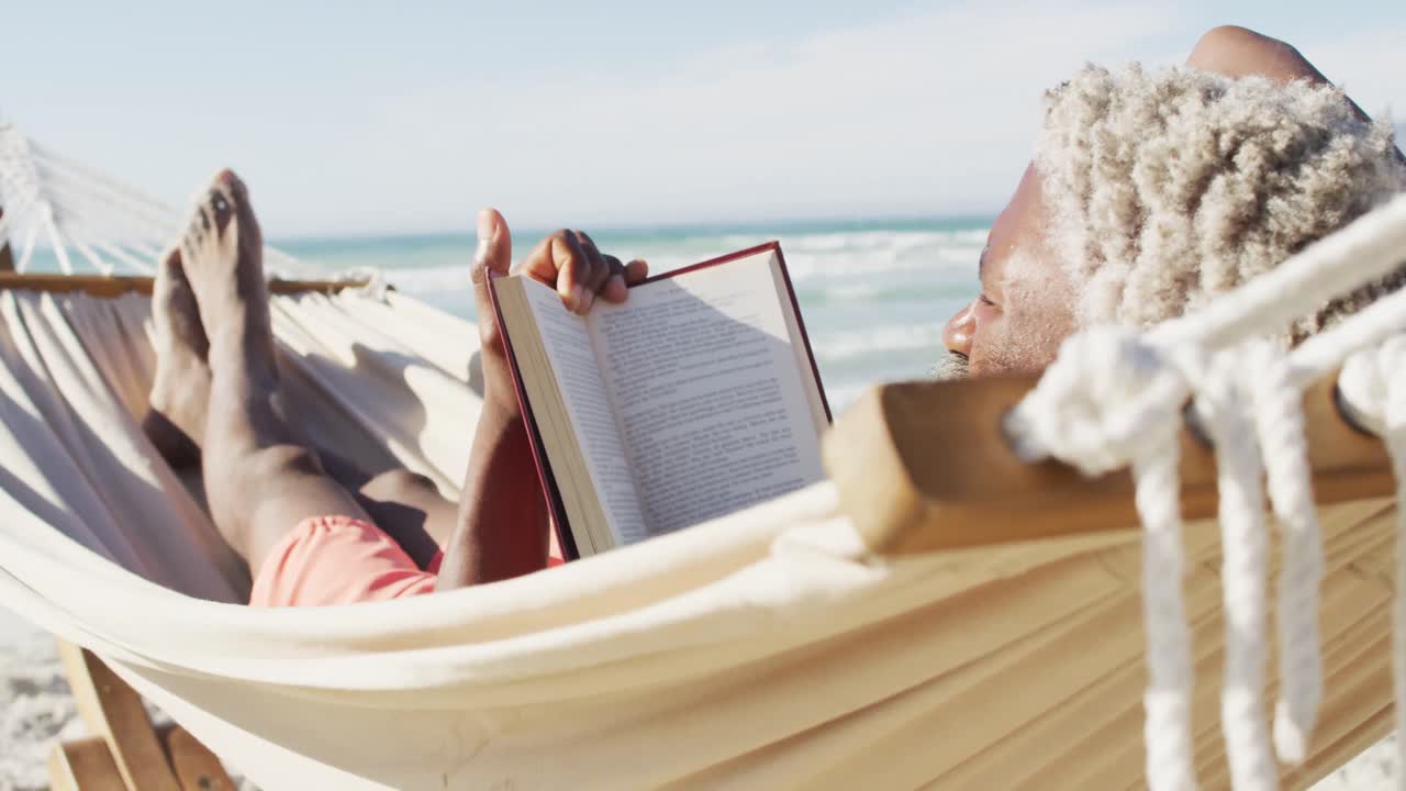 Senior african american man reading and lying in hammock on sunny beach