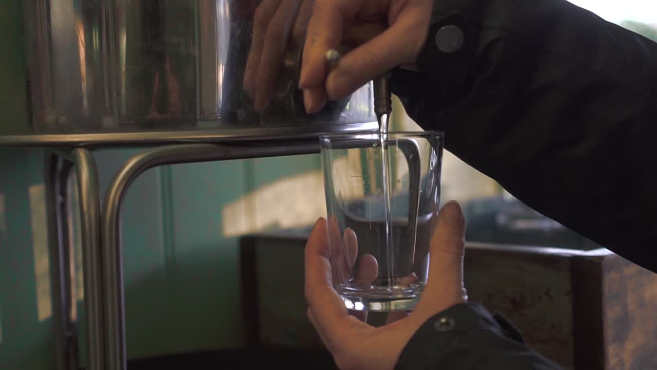 Girl water pours into the glass from a bigger metal container in slow motion.