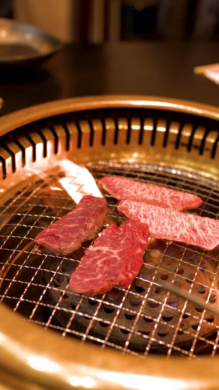 Primo piano di carne di manzo marmorizzata che cuoce su una griglia metallica rotonda con fiamme arancioni tremolanti.