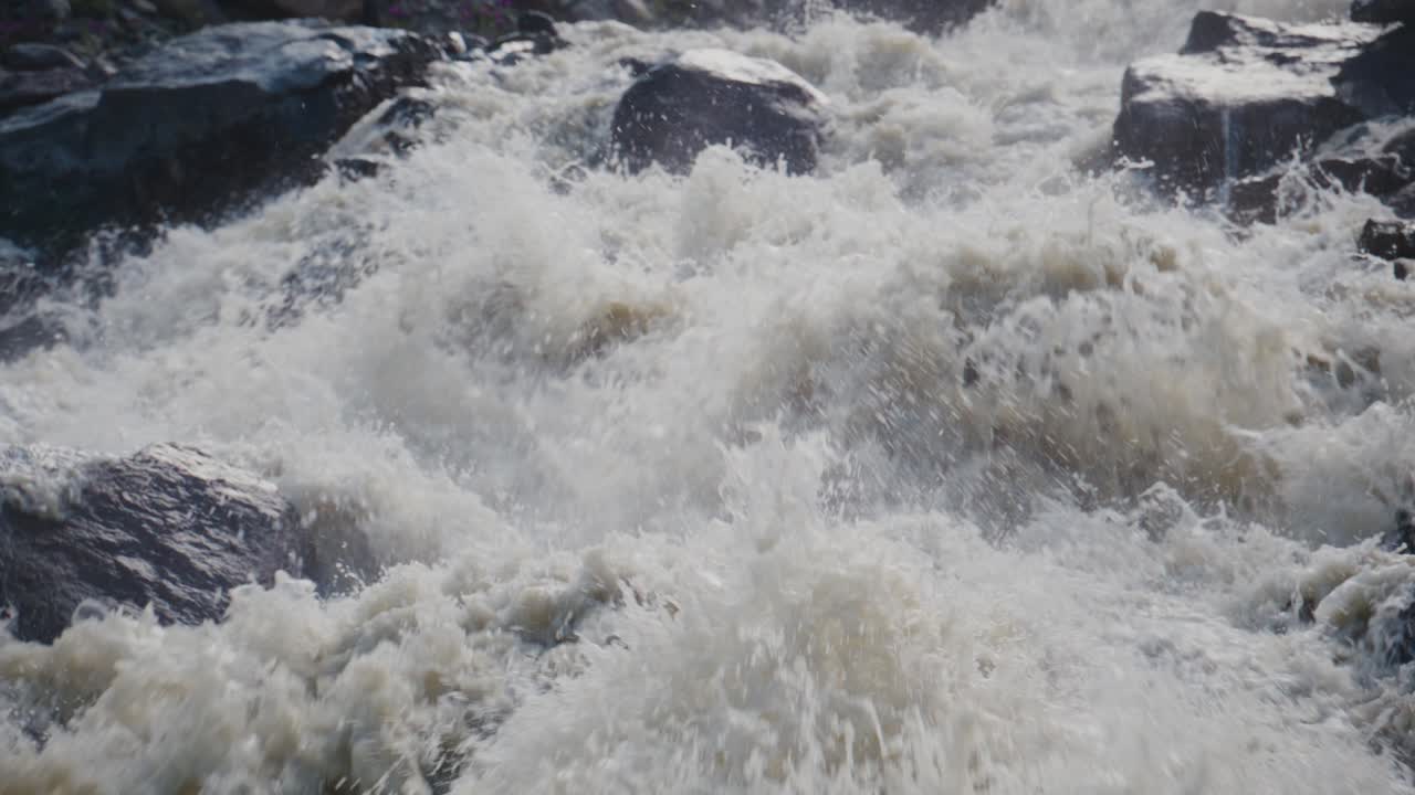 Raging river crashes over dark rocks. Foam explodes in chaotic white bursts. Water rushes with raw power and speed. Scene shows untamed nature's force. Ideal for adventure, danger