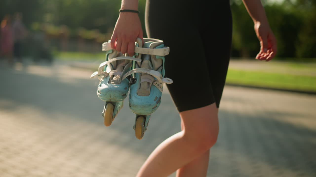 Lady wearing black tights walking outdoors holding cyan roller skates in her right hand on bright sunny day, background features lush greenery and blurred figures of people walking