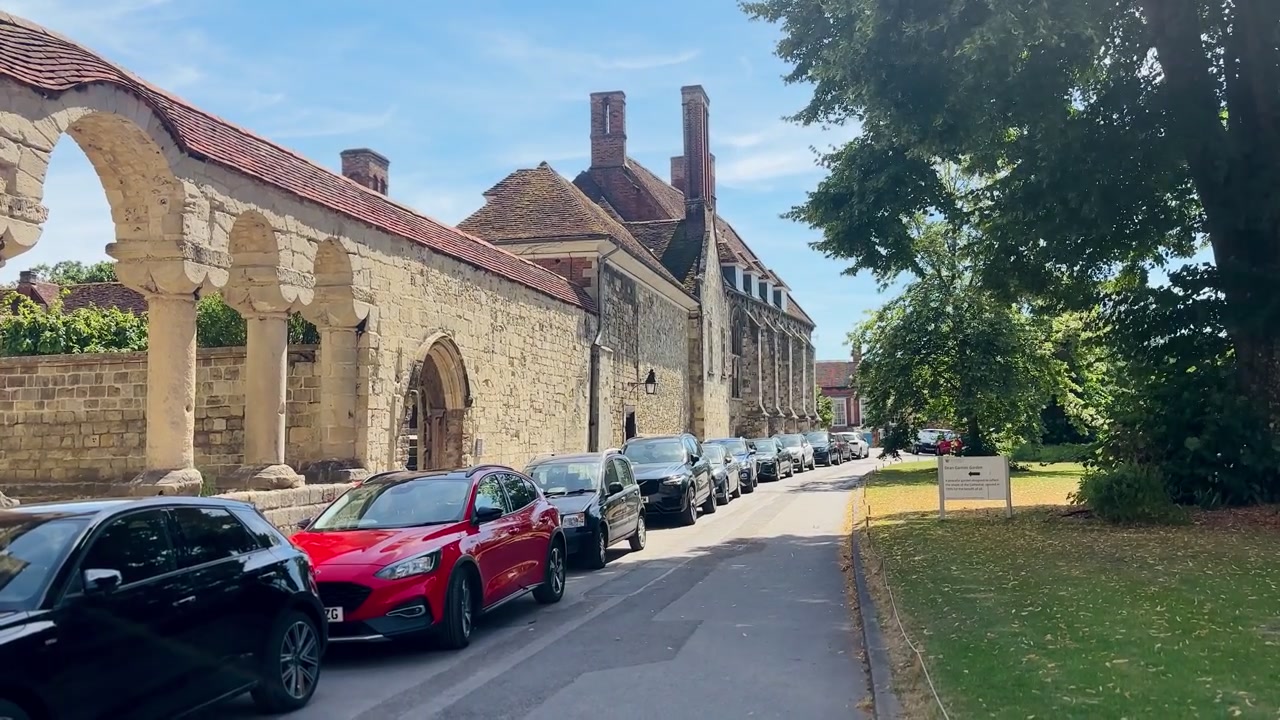 Via storica accanto al Cathedral Close con un porticato medievale in pietra, auto parcheggiate e alberi secolari a Winchester, Hampshire, Inghilterra, sotto un cielo azzurro.