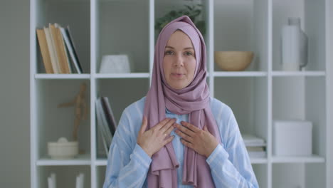 Looking-at-the-camera-muslim-girl-calling-friends-from-apartments-using-a-video-link-waving-with-hand-sitting-on-a-sofa-in-a-stylish-room-in-front-of-computer-screen-having-video-conversation