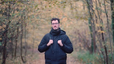 Alegre-Joven-Caballero-Alemán-Con-Gafas-Y-Barba,-Se-Encuentra-En-Medio-De-Los-Tonos-De-Un-Bosque-Mixto-Otoñal-Europeo,-Sonriendo-Cálidamente-Y-Ofreciendo-Un-Saludo-Amistoso