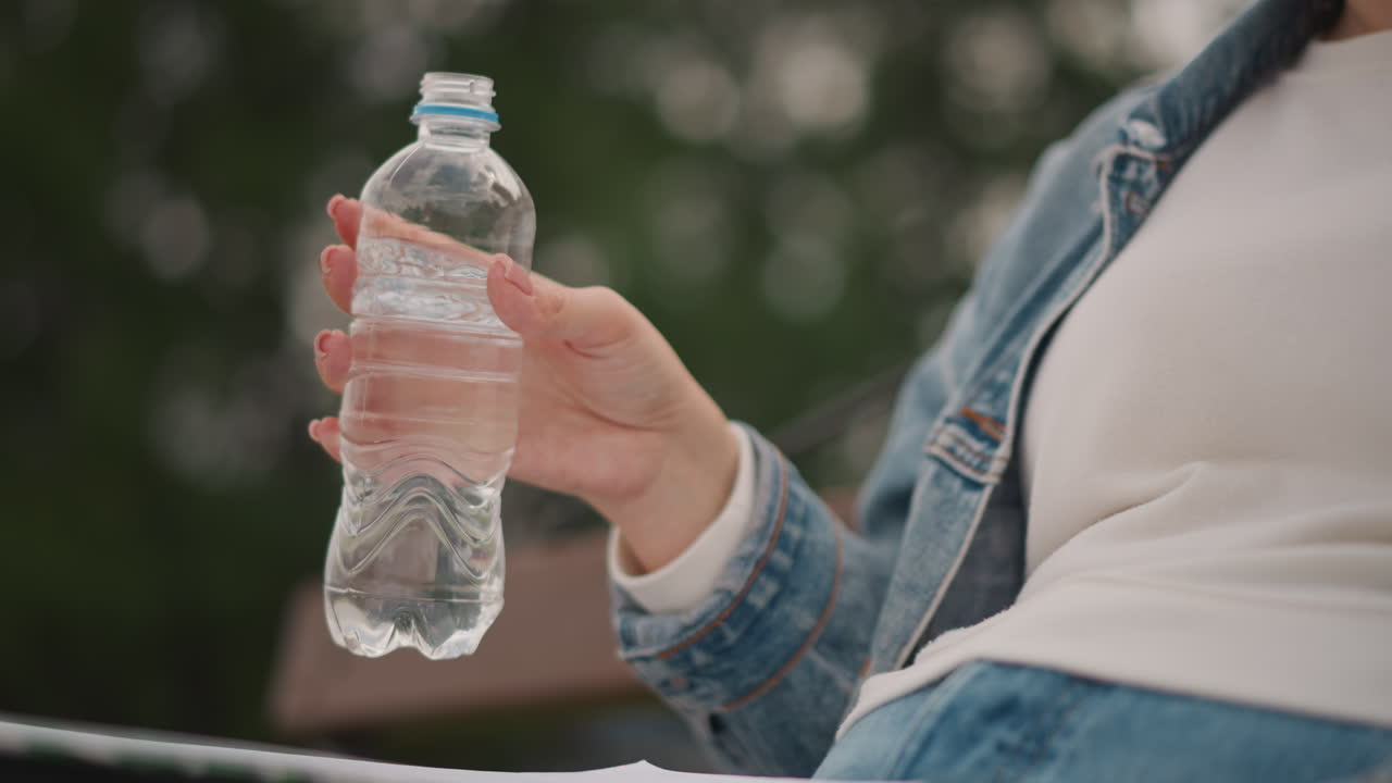 Naisen vartalokuva, joka pitää vesipulloa istuessaan penkillä, farkkutakki ja valkoinen t-paita näkyvissä, pehmeä bokeh-tausta, kondenssivettä pullossa, rento tauko kävelyn aikana, luonnonvalo.