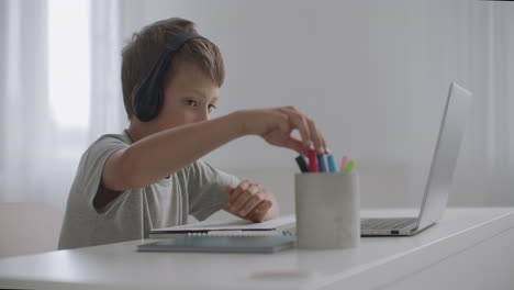 el escolar está descansando en su habitación escuchando música con auriculares conectados a una computadora portátil y coloreando imágenes en papel