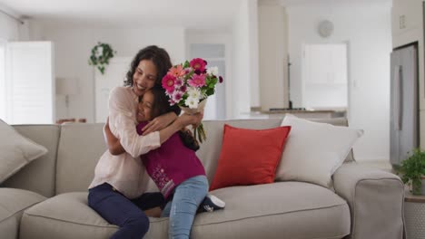 Happy-hispanic-woman-sitting-on-sofa-getting-flowers-from-daughter