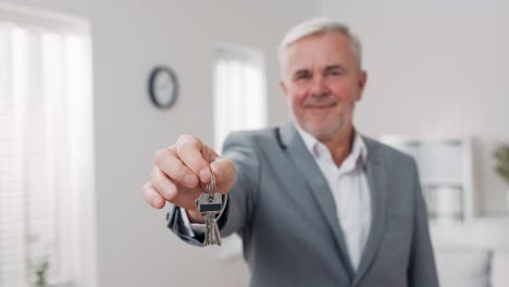 shot of apartment keys extended on palm of hand toward camera, gray-haired mature man dressed in suit hands over keys, real estate agent, owner of apartment, gives room to use